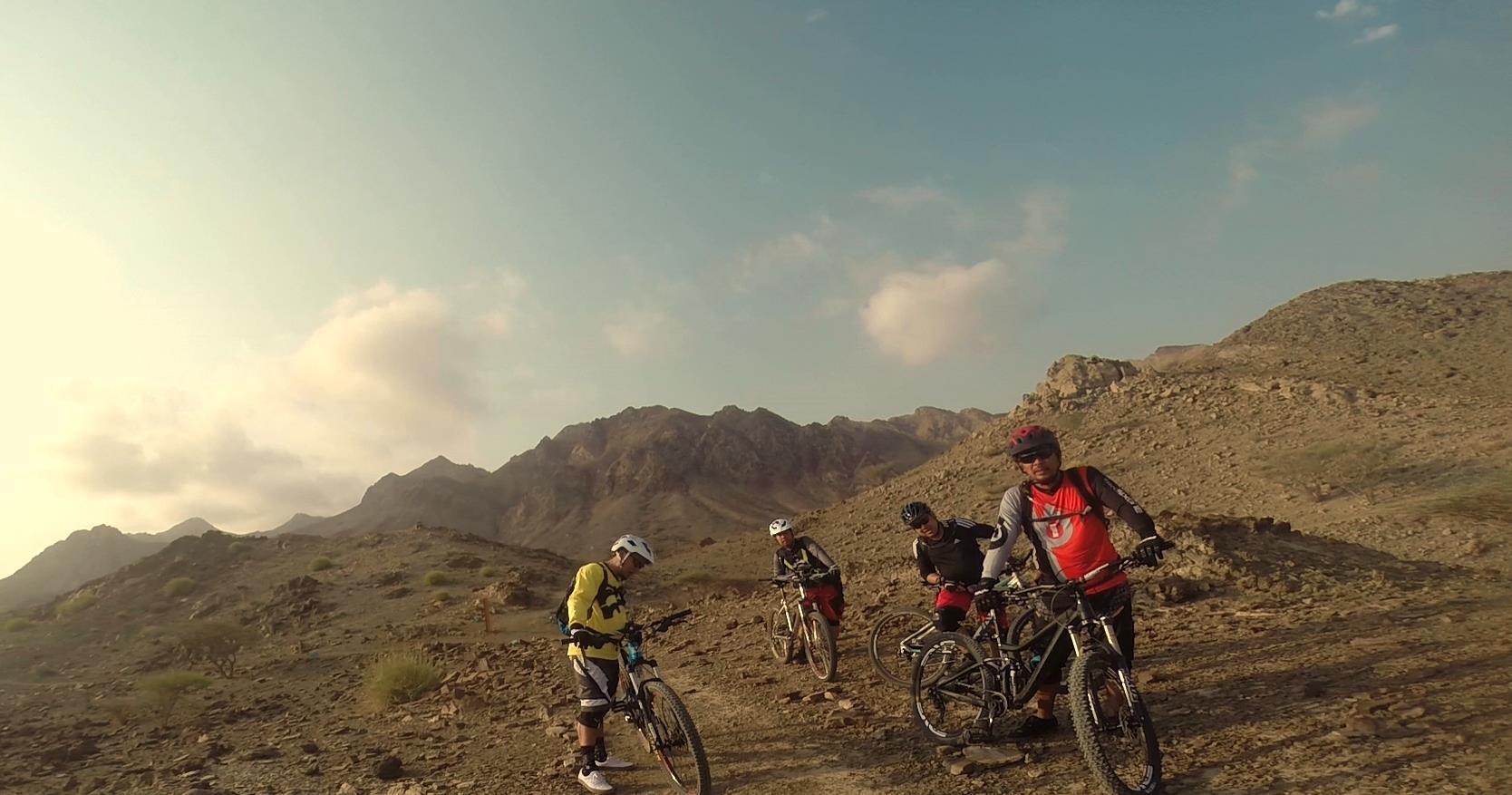 Four mountain bikers pause on a rocky trail in a desert landscape, surrounded by mountains under a partly cloudy sky. The riders are wearing protective gear and helmets, and they appear to be checking their bikes or planning their route. The terrain is rugged, with sparse vegetation. Showka Trails mountain bike trail.