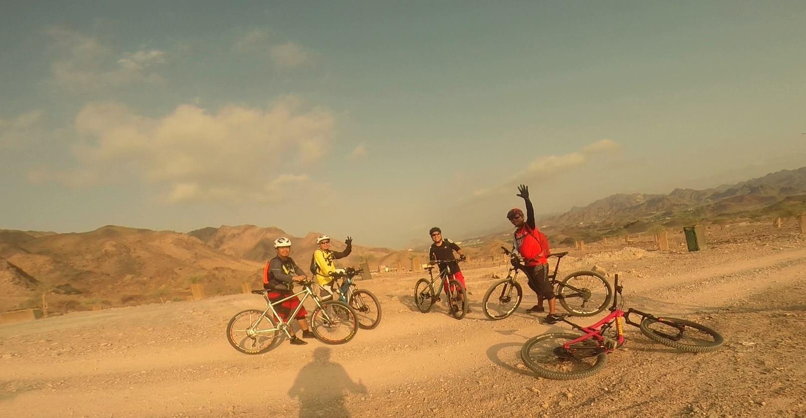 A group of five mountain bikers, wearing helmets and riding gear, stands on a dirt path surrounded by mountainous terrain. Some of them are waving and smiling at the camera, while a couple of bikes lie on the ground nearby. The sky is partly cloudy and the scene captures a sense of camaraderie and adventure in an outdoor setting. Showka Trails mountain bike trail.