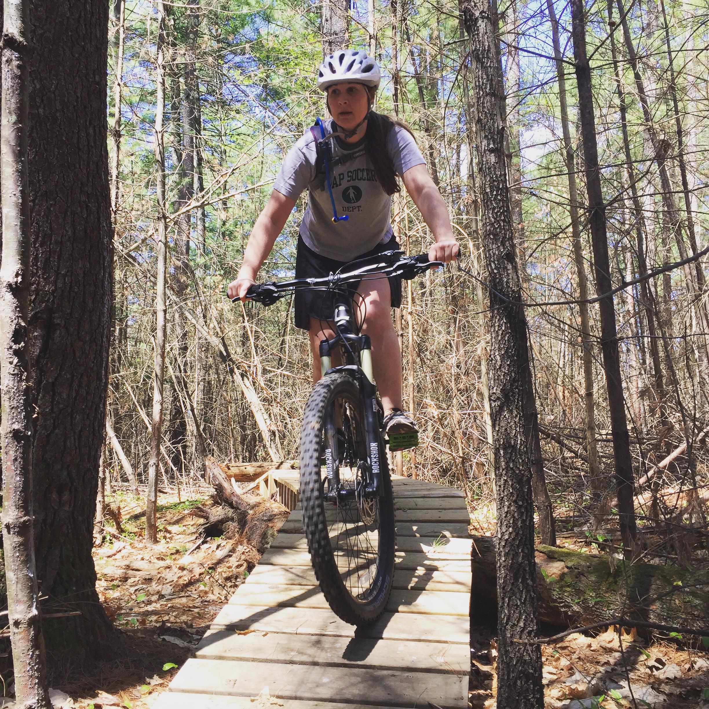 A person riding a mountain bike along a wooden bridge in a forested area, surrounded by trees and underbrush. The rider is wearing a helmet, a t-shirt, and shorts, displaying focus as they navigate the trail. Yudicky Farm mountain bike trail.