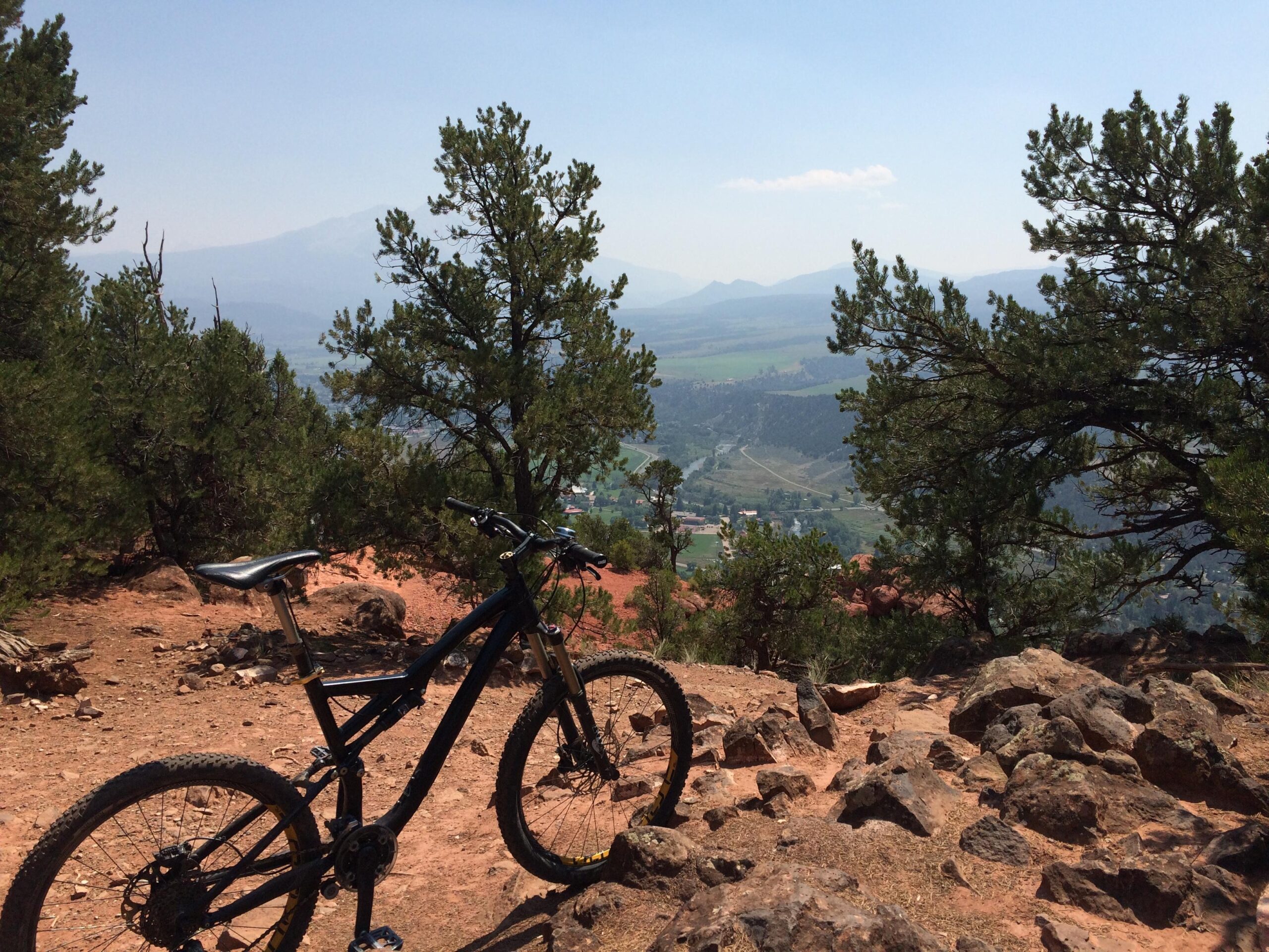 Specialized Stumpjumper FSR Expert Evo: A mountain bike rests on a rocky trail overlooking a valley filled with greenery and distant mountains under a clear blue sky. Surrounding vegetation includes pine trees, creating a scenic outdoor environment for cycling.