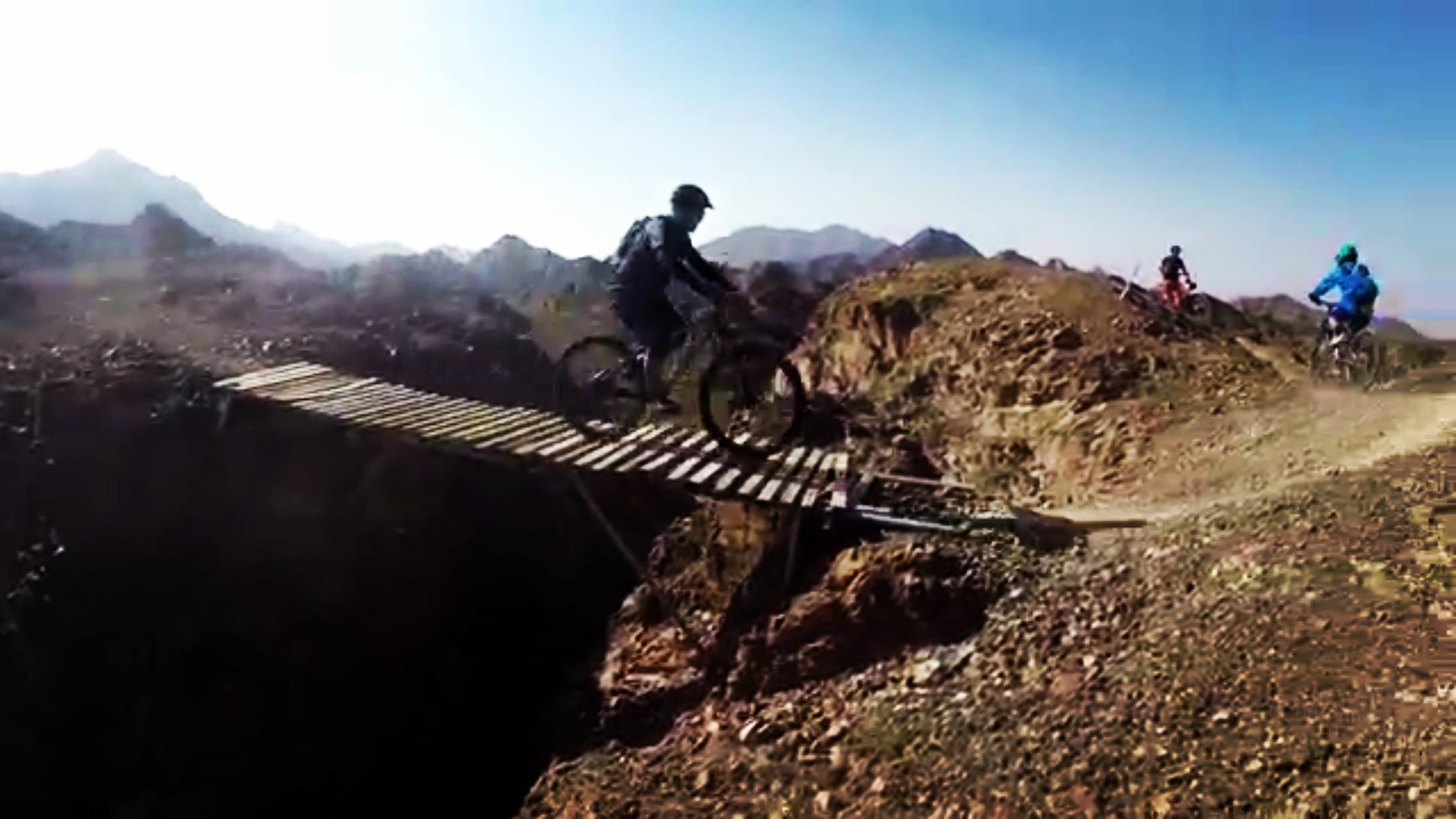 A mountain biker traverses a narrow wooden bridge over a steep drop, surrounded by rocky terrain and mountains in the background. The sky is clear and blue, suggesting a bright day for outdoor adventure. Two other cyclists can be seen in the distance, emphasizing the rugged landscape and the excitement of mountain biking. Showka Trails mountain bike trail.