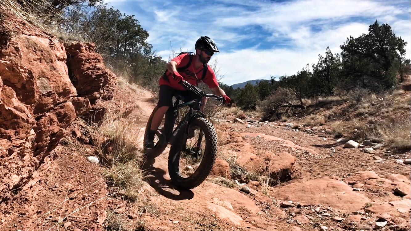 A person riding a fat bike on a rocky dirt trail surrounded by red dirt, grass, and sparse vegetation under a blue sky with scattered clouds. The rider is wearing a helmet and protective gear, showcasing an action-packed moment as they navigate the rugged terrain. Golden Open Space mountain bike trail.