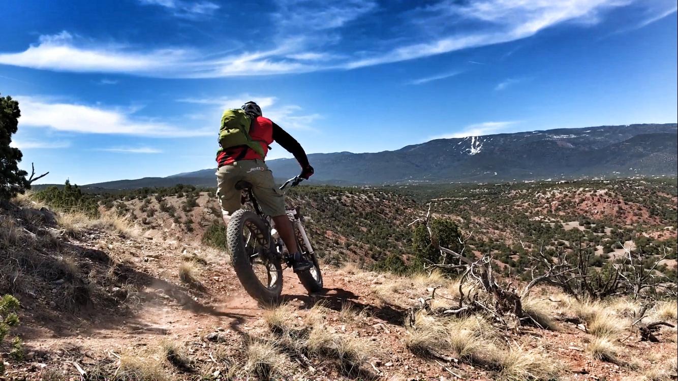 A person riding a mountain bike on a rocky trail, with a panoramic view of rolling hills and mountains under a blue sky. The cyclist is wearing a red shirt and a green backpack, navigating a downhill path surrounded by desert vegetation. Dust kicks up from the bike tires as they descend. Golden Open Space mountain bike trail.
