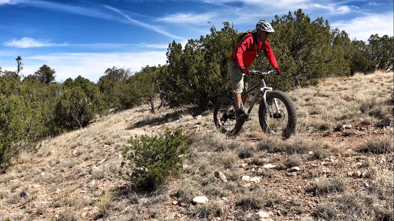 A mountain biker in a red jacket and helmet rides a fat bike on a rocky trail surrounded by shrubbery and grasses under a blue sky. Golden Open Space mountain bike trail.