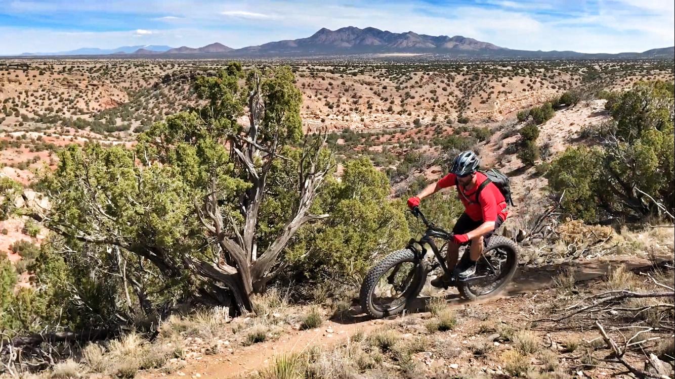 A mountain biker riding a fat bike on a dirt trail, surrounded by scenic desert hills and scrub vegetation, with distant mountains under a blue sky. Golden Open Space mountain bike trail.