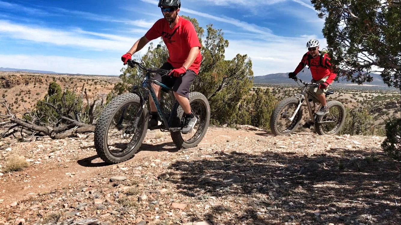 Two mountain bikers riding on a rocky trail surrounded by shrubs and a distant landscape under a blue sky. Both riders are wearing helmets and red shirts, with one on a black bike and the other on a lighter-colored bike. The terrain is rugged and offers a scenic view of the surrounding area. Golden Open Space mountain bike trail.