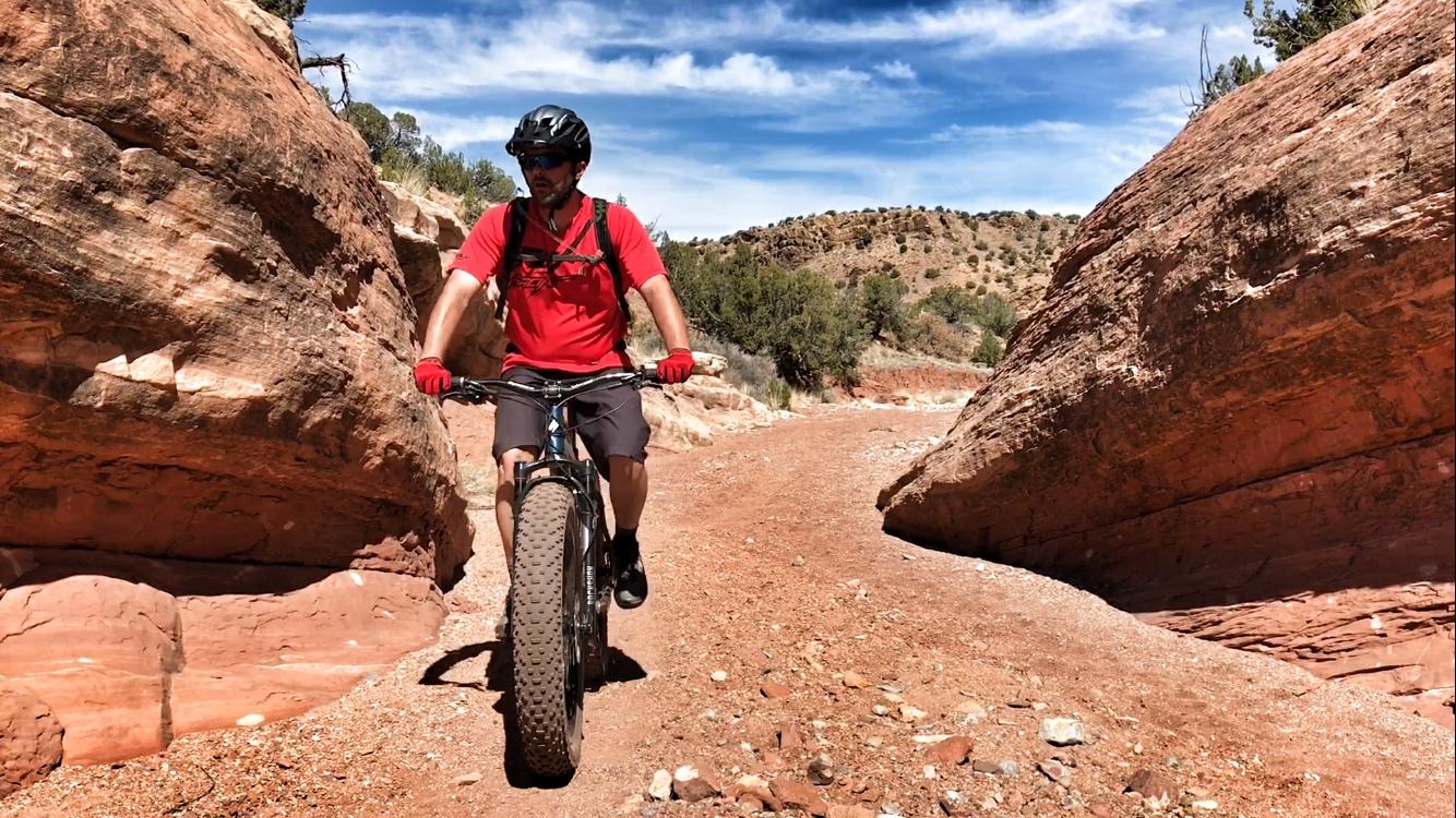 A person riding a fat tire bike on a rocky trail between towering red rock formations, with a blue sky and distant hills visible in the background. The cyclist is wearing a helmet and a bright red shirt, showcasing an adventurous outdoor scene. Golden Open Space mountain bike trail.