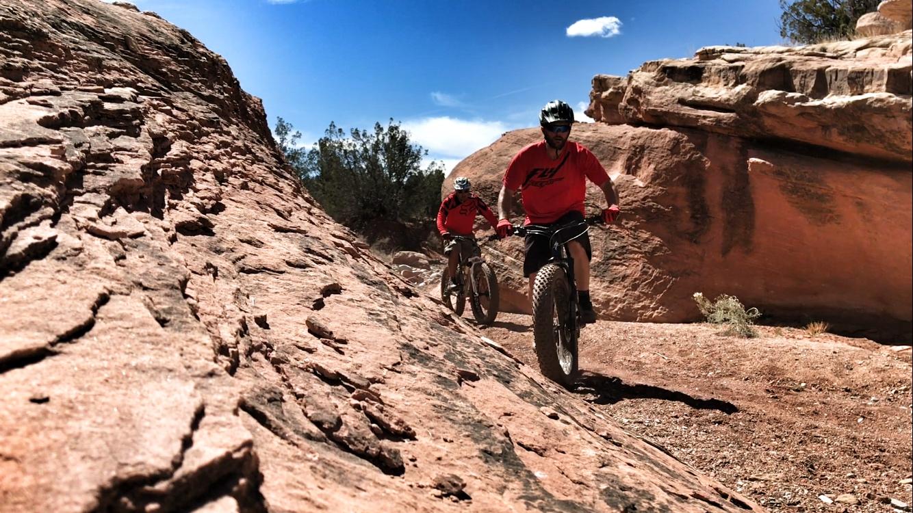 Two mountain bikers ride along a rocky terrain under a clear blue sky. The bikers, wearing helmets and red jerseys, navigate a sloped, reddish landscape with boulders and sparse vegetation in the background. Golden Open Space mountain bike trail.