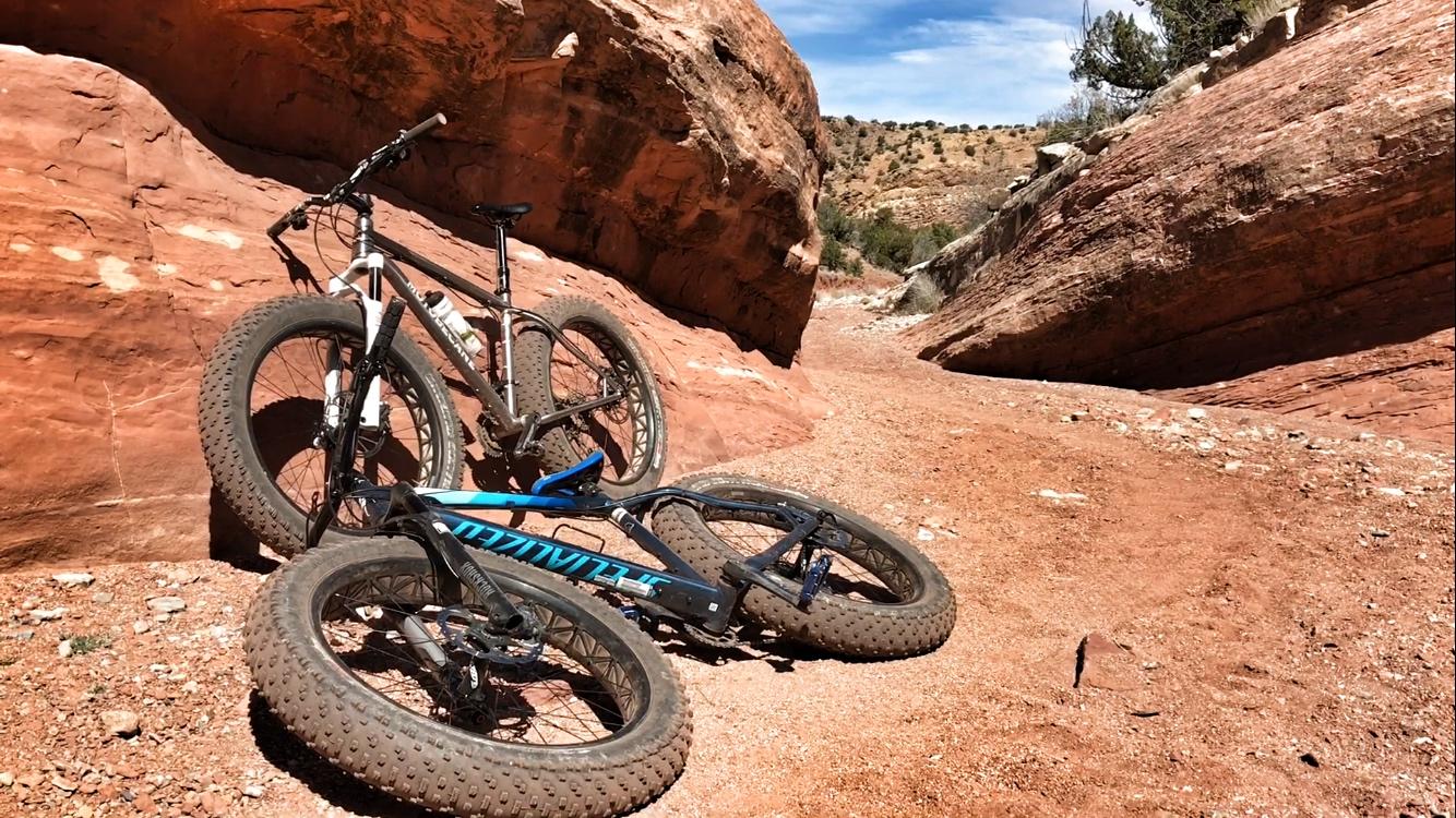 Two mountain bikes are leaning against a red rock wall in a canyon-like terrain, surrounded by a sandy path. The bikes have thick tires, suitable for off-road riding, and the landscape features rocky outcrops and sparse vegetation under a clear blue sky. Golden Open Space mountain bike trail.