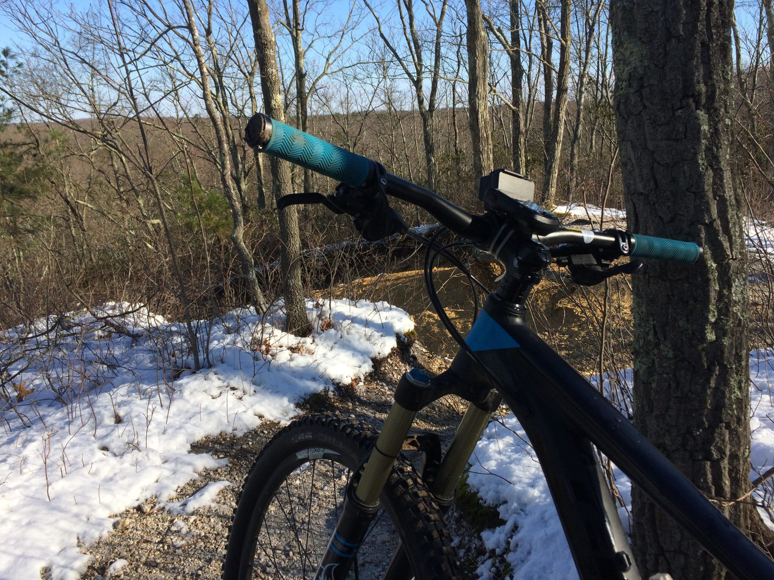 Close-up view of a mountain bike handlebars with turquoise grips, set against a winter landscape featuring patches of snow and bare trees. The background shows a path through the woods, indicating a tranquil outdoor scene. Allaire State Park mountain bike trail.