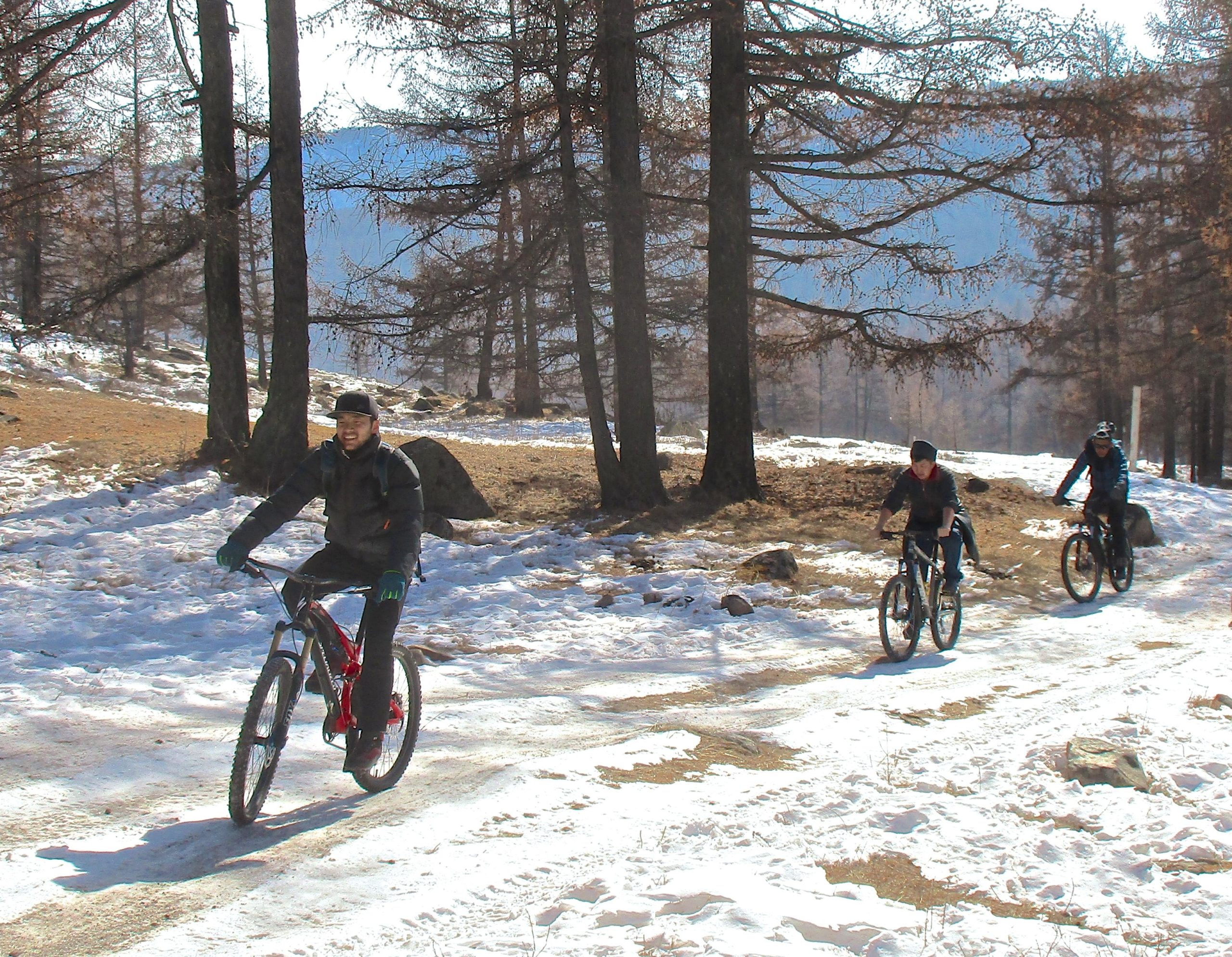 Four cyclists riding mountain bikes along a snowy trail surrounded by trees on a sunny winter day. The first rider is smiling and wearing a black jacket and gloves, while the others are focused on their ride in the background. Snow covers part of the ground, creating a contrasting landscape with patches of dirt and grass. Tsetsee Gun East Trail mountain bike trail.