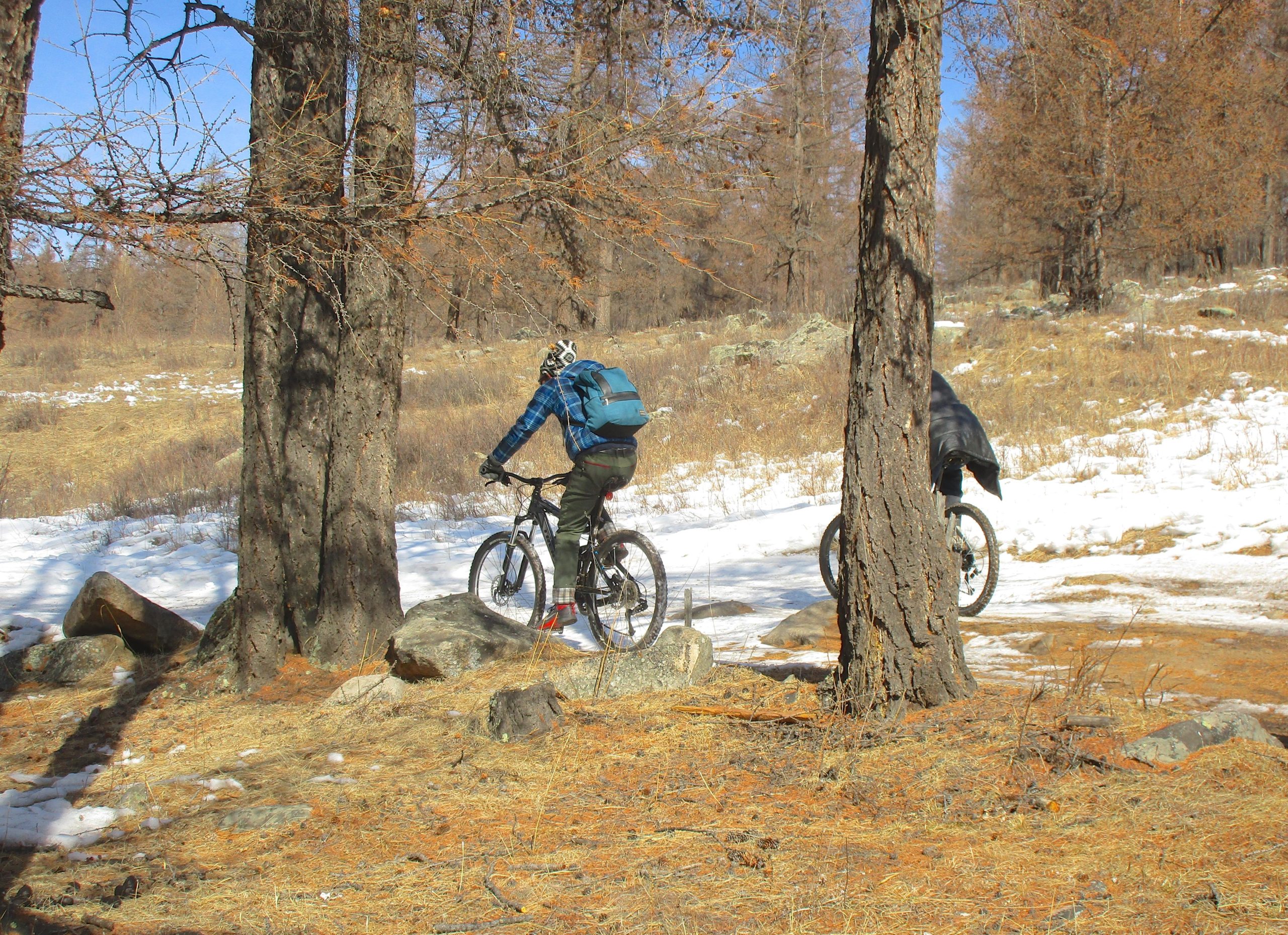 Two mountain bikers riding on a snow-covered path through a forest, surrounded by tall trees and rocky terrain. The scene features a mix of dry grass and patches of snow, indicating early spring conditions. One biker is wearing a blue plaid jacket and a backpack, while the other is dressed in darker clothing and slightly hidden behind a tree. Tsetsee Gun East Trail mountain bike trail.