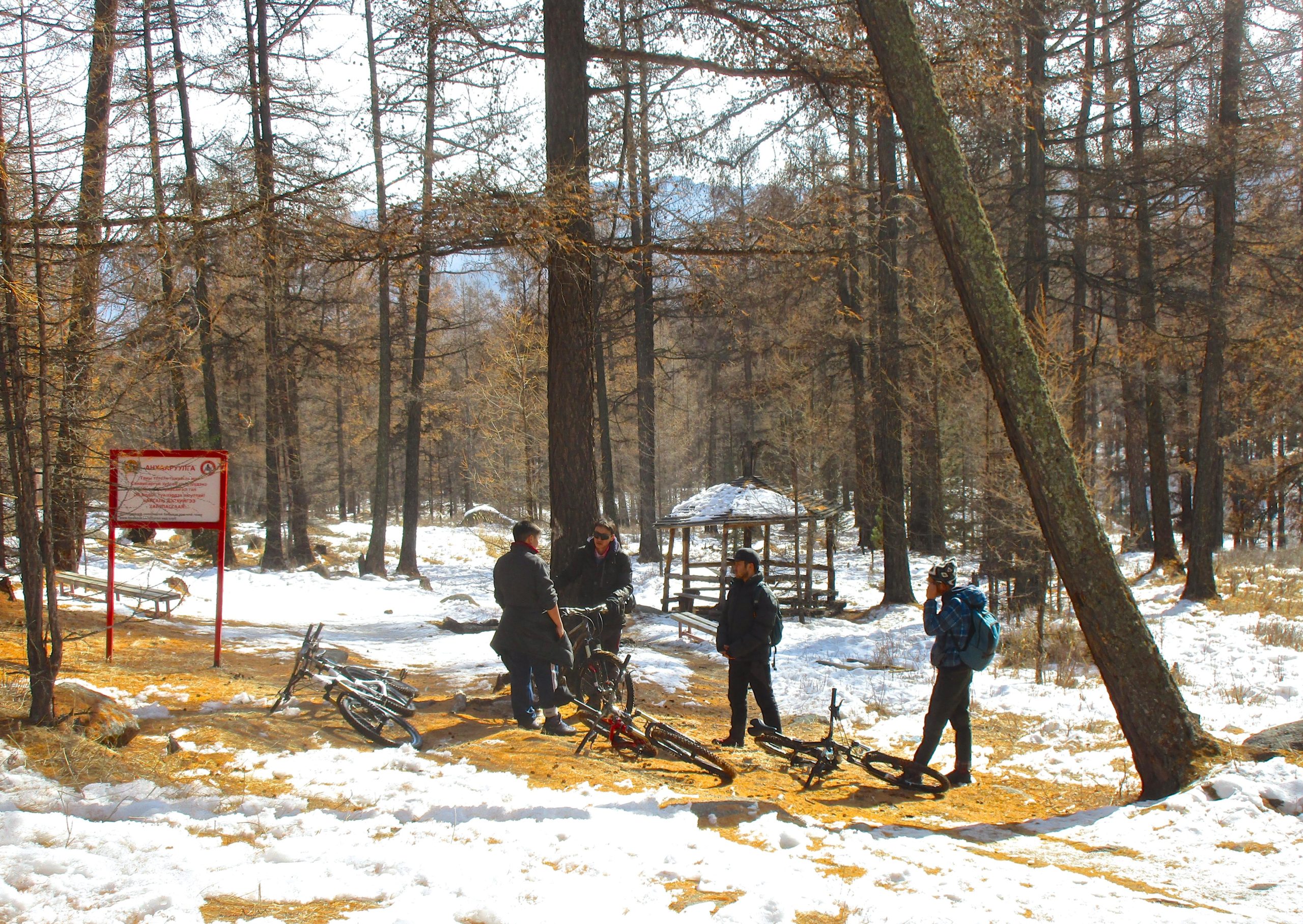 A group of four individuals standing near bicycles in a snowy forest area. They are engaged in conversation, with a wooden gazebo visible in the background along with a signpost. The ground is a mix of snow and earthy tones, and tall trees surround the scene. Tsetsee Gun East Trail mountain bike trail.