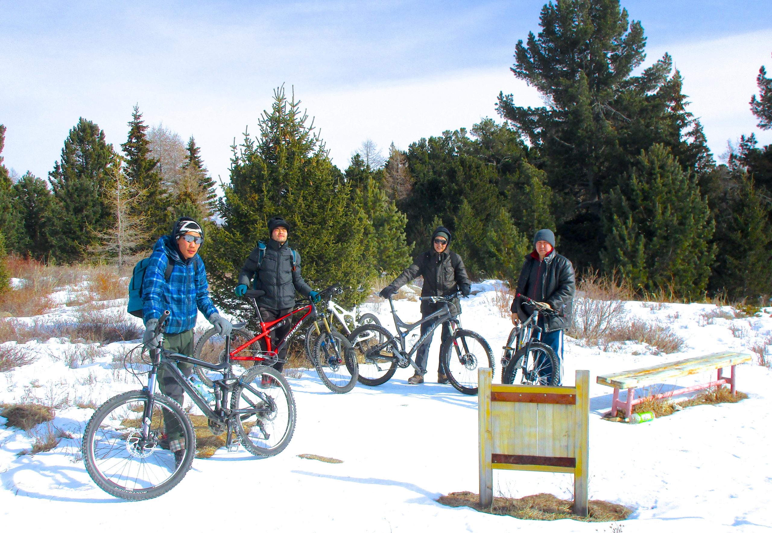 Four mountain bikers are standing on a snowy trail in a forested area. They are dressed in winter clothing, with two of them wearing jackets and hats, while others wear flannel and sunglasses. Each biker has a different style of bike. In the background, there are evergreen trees and a clear sky. A wooden sign and a bench are visible in the foreground. Tsetsee Gun East Trail mountain bike trail.