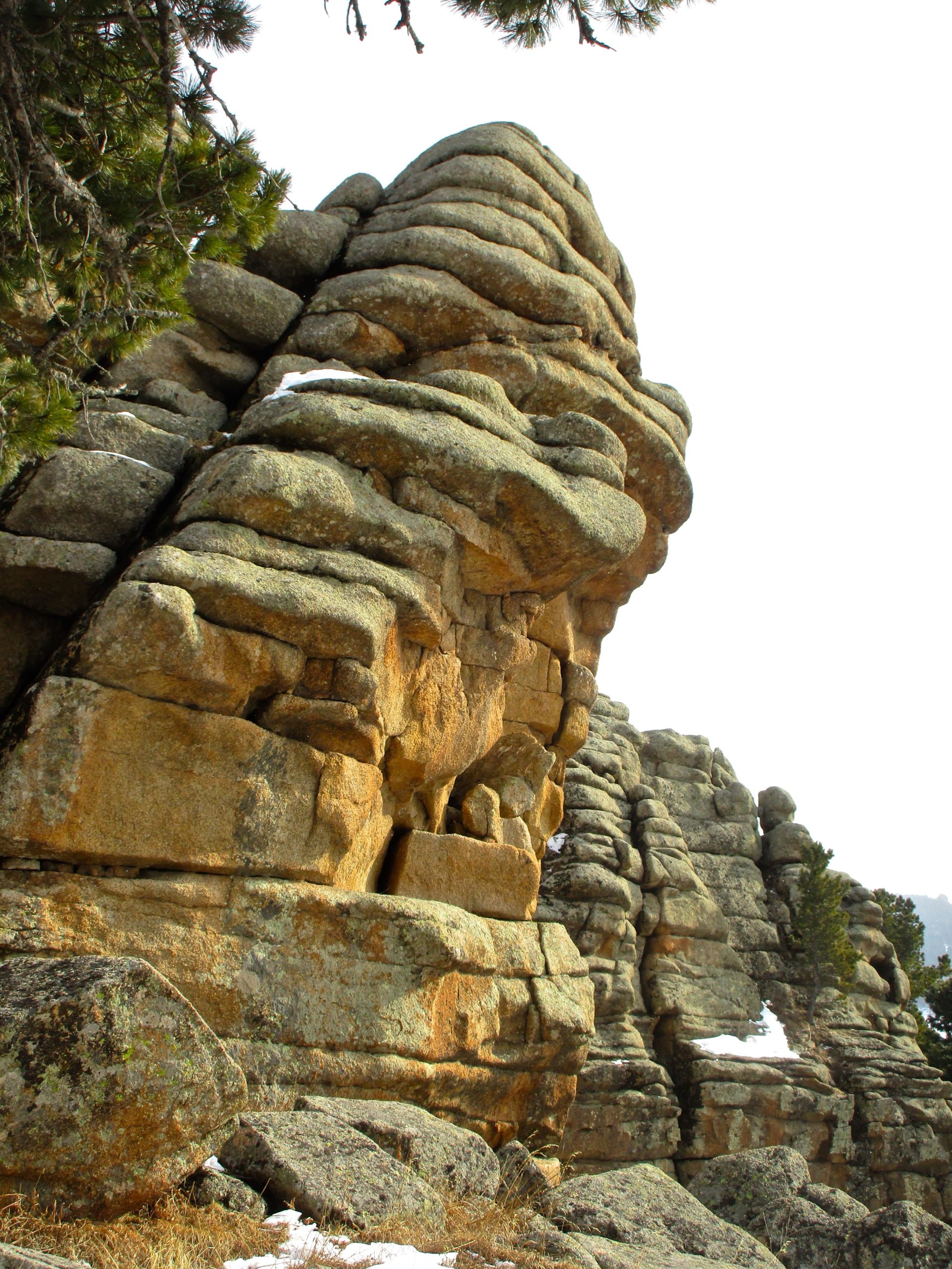An angular rock formation with layered, textured surfaces, set against a bright sky. Sparse pine trees are visible near the top, and patches of snow can be seen on the rocks and surrounding ground. The scene conveys a rugged natural landscape. Tsetsee Gun East Trail mountain bike trail.