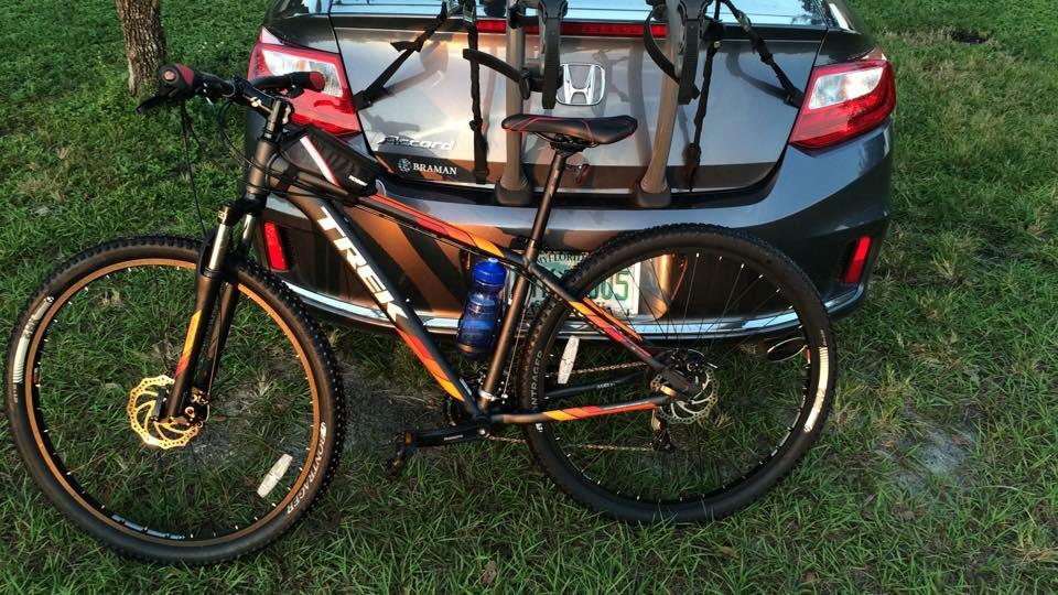 Trek Marlin 5: A mountain bike is secured to a bike rack on the back of a gray Honda vehicle, parked on grassy ground. A water bottle is attached to the bike frame. The scene is illuminated by soft sunlight.