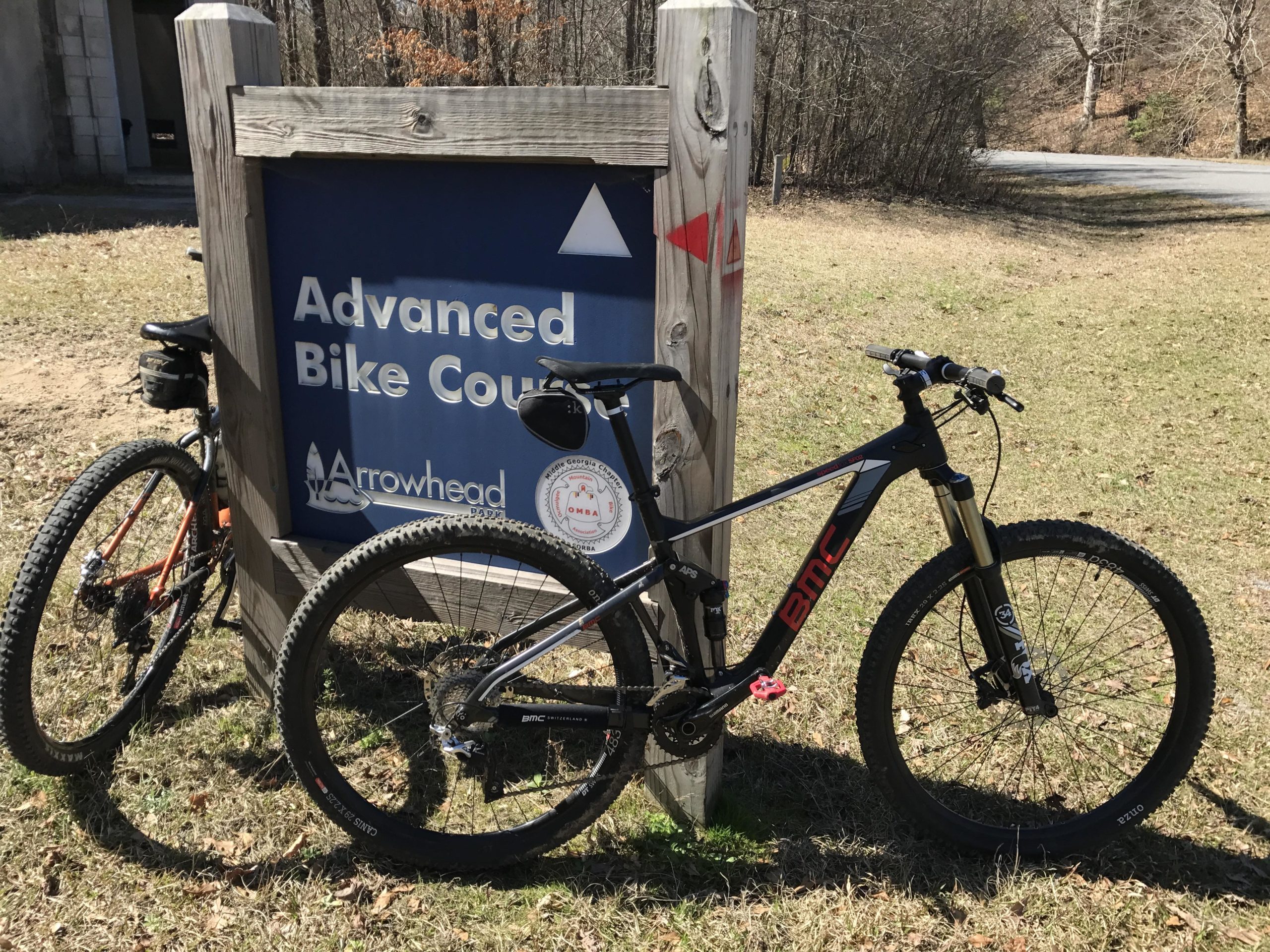 Image of two mountain bikes parked beside a sign that reads "Advanced Bike Course" at Arrowhead Park. The sign indicates the trail designation with an upward triangle symbol. The scene is set in a natural outdoor environment with grass and trees visible in the background. Arrowhead Park mountain bike trail.