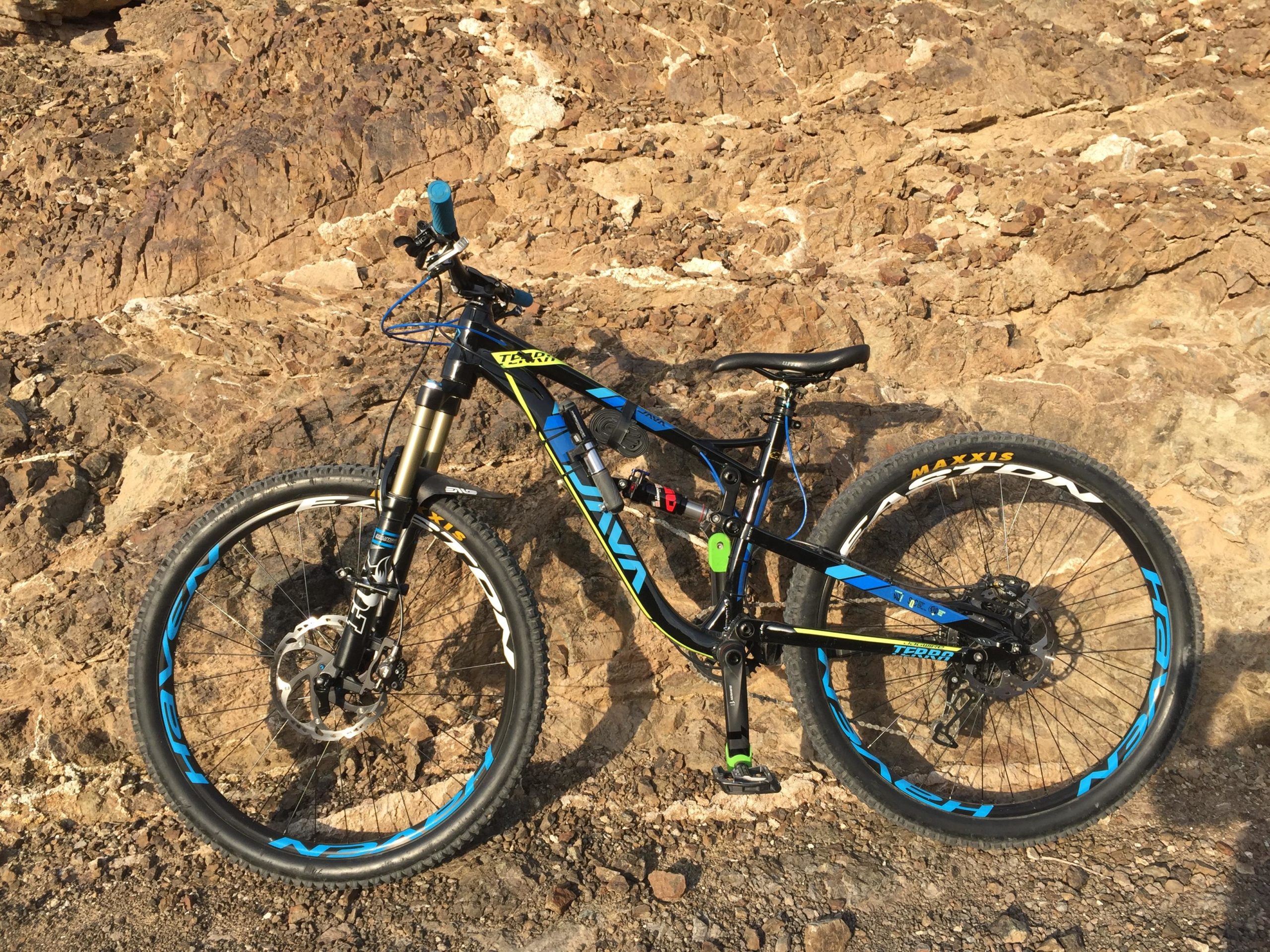A blue and yellow mountain bike leaning against a rocky background, showcasing its tires and frame details on a gravel path. Showka Trails mountain bike trail.