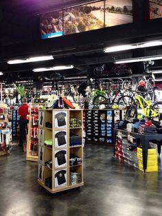 An indoor retail store showcasing a variety of bicycles and sporting goods. In the foreground, a wooden display rack contains t-shirts, while the background features rows of bicycles and athletic gear. Bright lighting illuminates the space, and large screens with outdoor imagery are visible on the walls.