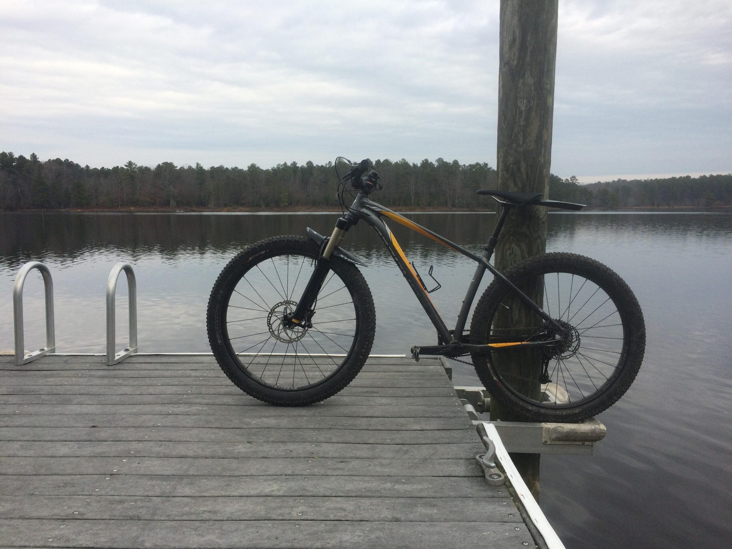 Specialized Fuse Comp 6Fattie: A mountain bike is positioned on a wooden dock extending over a calm lake. The background features a forest of trees lining the shoreline under a cloudy sky.