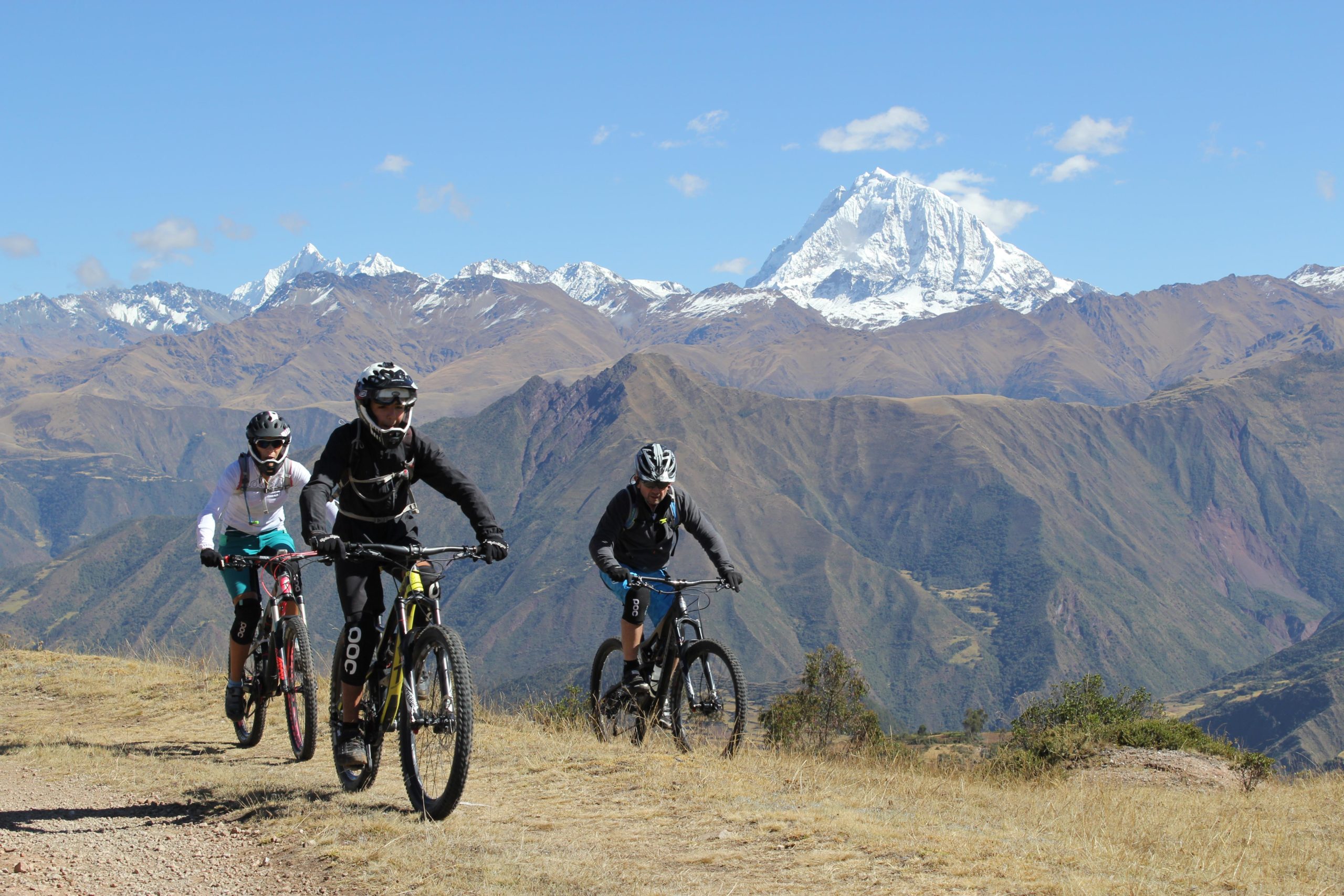 Three mountain bikers navigating a rocky trail with expansive views of mountain ranges and snow-capped peaks in the background. The landscape features golden grasses and rugged terrain under a clear blue sky. Limatambo DH mountain bike trail.
