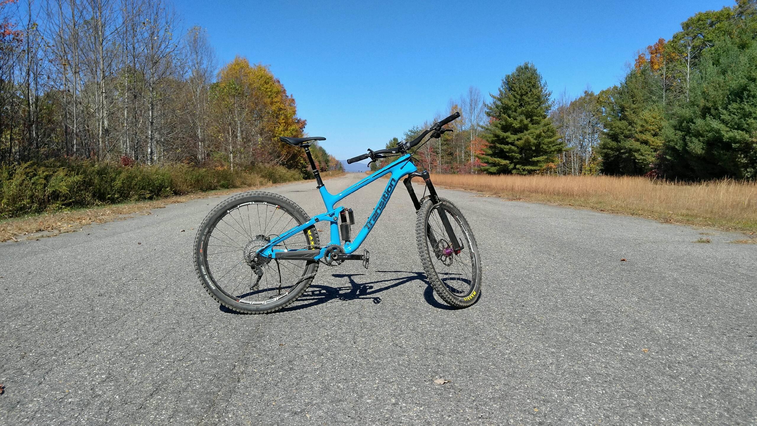 Transition Patrol: A blue mountain bike positioned on a gravel road surrounded by autumn foliage and trees under a clear blue sky.