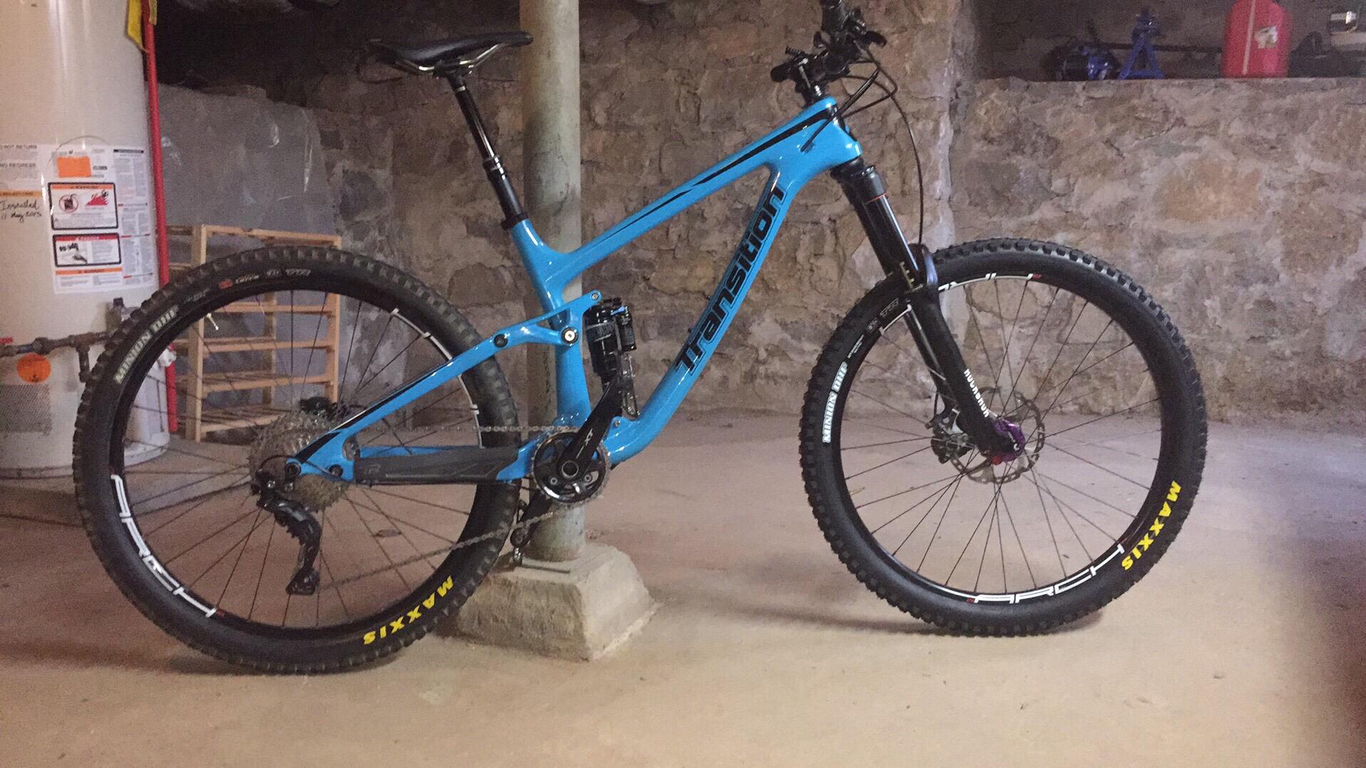 Transition Patrol: A blue mountain bike leaning against a concrete pillar in a basement with stone walls. The bike features large tires suitable for off-road terrain, a suspension system, and detailed components visible on the frame. The background includes a water heater and various tools.