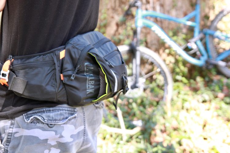A close-up view of a person wearing a black CamelBak hip pack, featuring orange accents, secured around the waist. In the background, a blue mountain bike is partially visible, surrounded by greenery. The individual's clothing includes a black shirt and camouflage-patterned shorts.