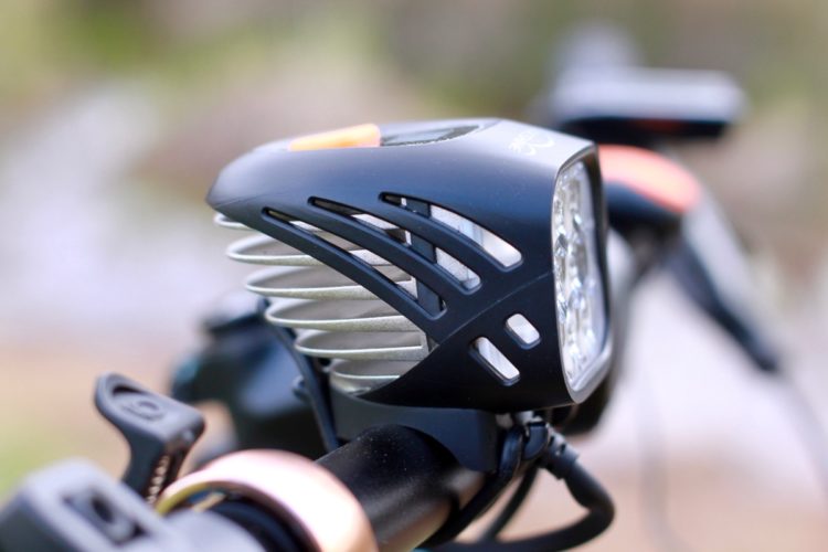 Close-up of a bicycle headlight mounted on handlebars, featuring a sleek black casing with ventilation slots and a bright LED light at the front. The background is blurred, emphasizing the headlight's design.