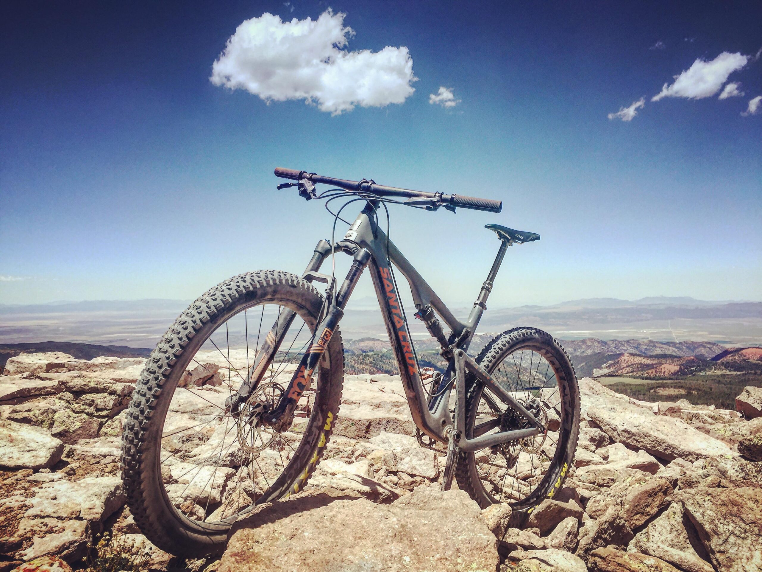 Santa Cruz Tallboy Carbon: A mountain bike positioned on rocky terrain, overlooking a vast landscape with mountains and a clear blue sky. The bike features stout tires and a sleek frame, emphasizing a sense of adventure and outdoor exploration.