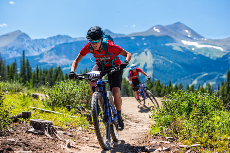 Two mountain bikers navigate a rugged trail in a mountainous landscape. The foreground features a cyclist wearing a red shirt and a blue helmet, focused on the path ahead. In the background, another rider extends the scene with scenic mountain peaks and greenery surrounding the trail under a clear blue sky.