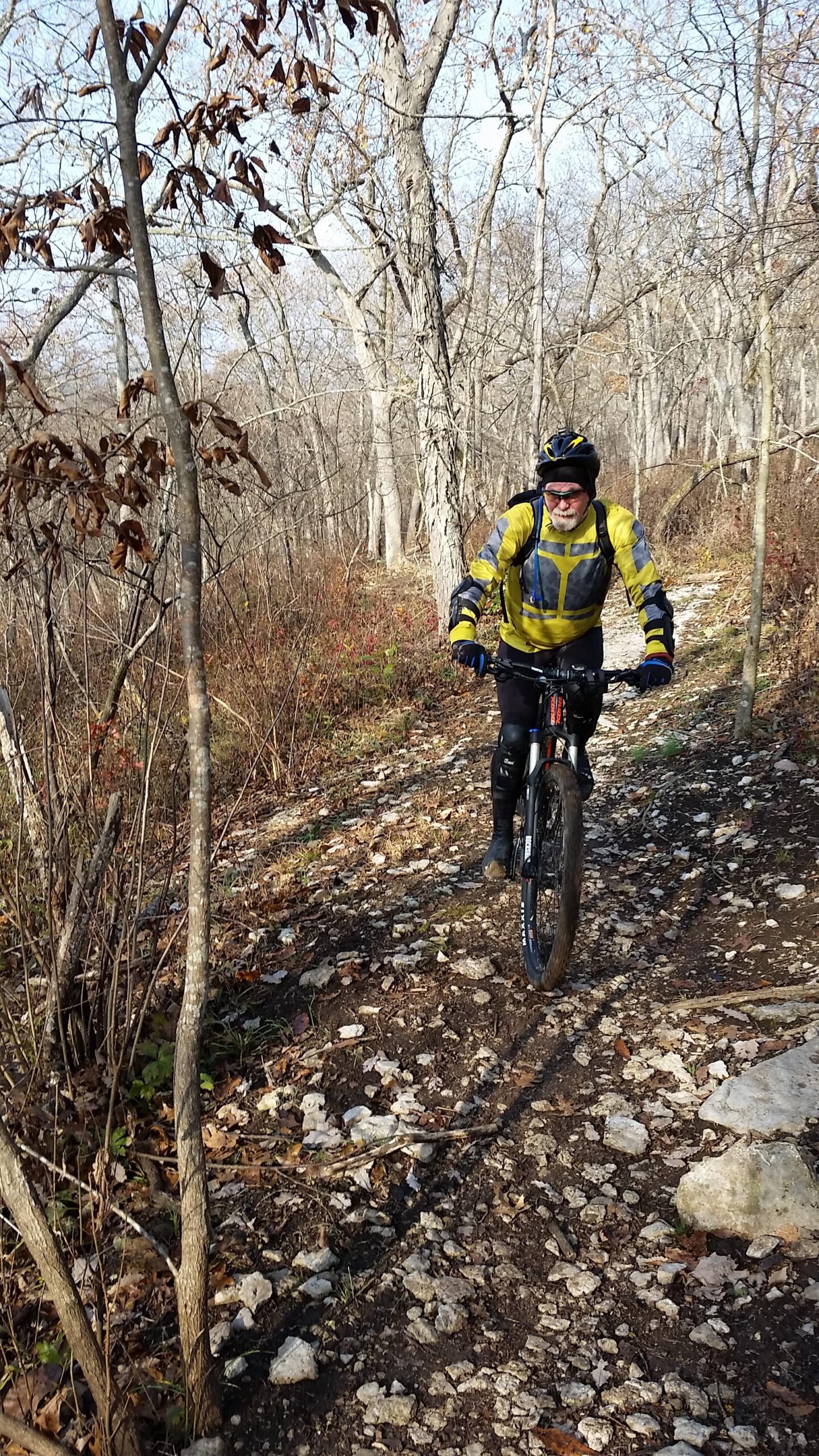 Kona Hei Hei: A person wearing a yellow and gray biking outfit, along with a helmet, is riding a mountain bike along a rocky trail in a wooded area. The surrounding trees are bare, indicating early winter, with some leaves scattered on the ground.