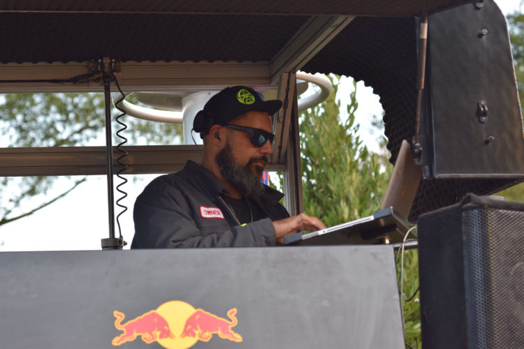 A DJ wearing sunglasses and a cap is focused on his laptop while performing in a mobile setup with an audio mixer. The background features trees and a Red Bull logo on the equipment, indicating a lively outdoor event.