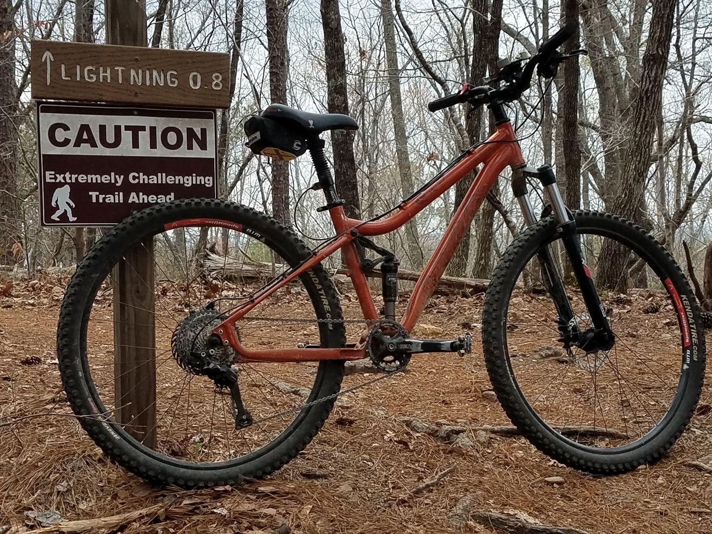 A mountain bike is parked next to a sign that indicates the "Lightning" trail, which is 0.8 miles ahead and marked as "Caution: Extremely Challenging Trail Ahead." The scene is set in a wooded area with bare trees and pine needles covering the ground. Oak Mountain State Park Bump Trail mountain bike trail.