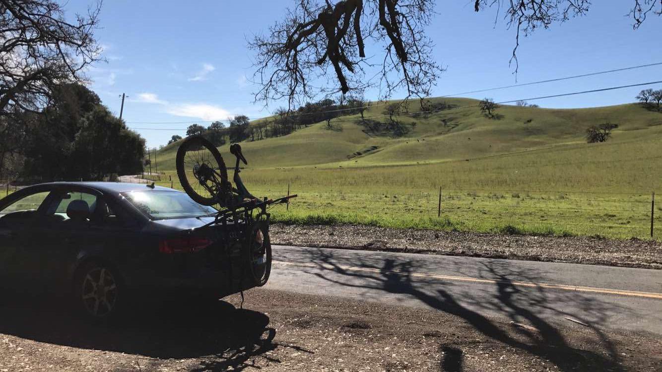 A parked car with a bicycle mounted on the rear rack, set against a backdrop of rolling green hills under a clear blue sky. The scene features a distant road, trees lining the landscape, and a peaceful rural atmosphere. Oyster Point mountain bike trail.
