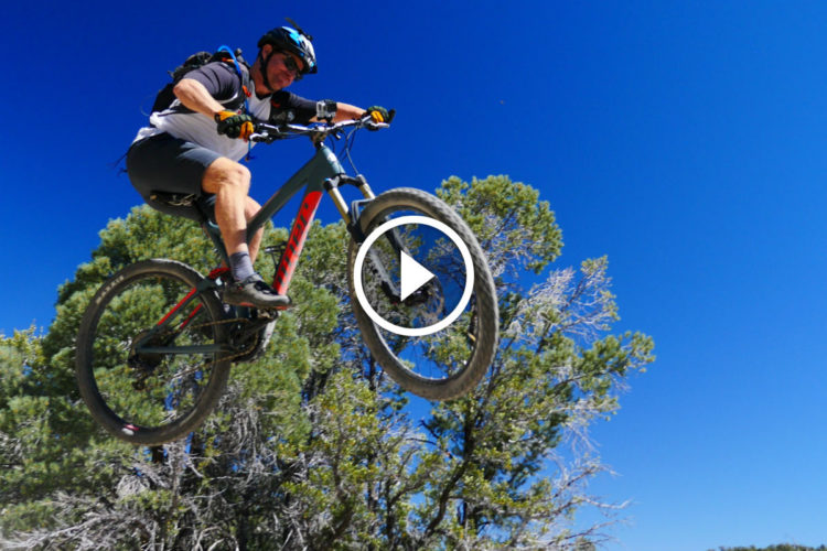 A mountain biker performing a mid-air jump over a trail, surrounded by trees, against a clear blue sky. The rider is wearing a helmet and protective gear, showcasing an action-packed moment in outdoor biking.