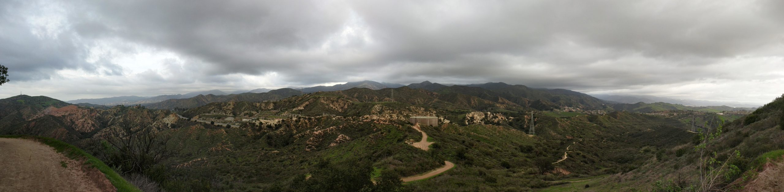 A panoramic view of rolling hills and mountains under a cloudy sky, with winding dirt paths and patches of greenery amongst rocky terrain. The landscape features varied elevations and a mix of light and shadow, capturing the natural beauty of the outdoors. Whiting Ranch Wilderness Park mountain bike trail.