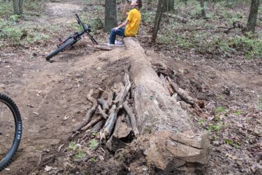 A person wearing a yellow shirt sits on a fallen log in a forested area, taking a drink from a green water bottle. A mountain bike rests nearby, and the ground is covered with leaves and branches, creating a natural setting for outdoor activities. Frost Woods mountain bike trail.