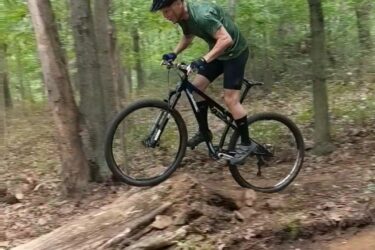 A mountain biker leaping over a log ramp on a dirt trail in a wooded area, surrounded by green trees and foliage. The cyclist wears a helmet and bike gear, demonstrating an active and adventurous spirit. Frost Woods mountain bike trail.
