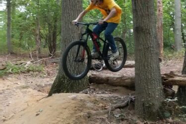 A young person in a yellow shirt and a blue helmet is riding a mountain bike over a small jump in a wooded area. The bike's front wheel is elevated off the ground as they navigate a trail surrounded by trees and scattered leaves. Frost Woods mountain bike trail.