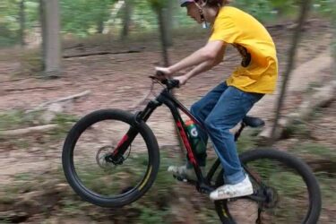 A person wearing a pink helmet and a yellow shirt is mid-air while riding a mountain bike over a log in a wooded area. The background features green trees and a dirt path, emphasizing an outdoor cycling activity. Frost Woods mountain bike trail.