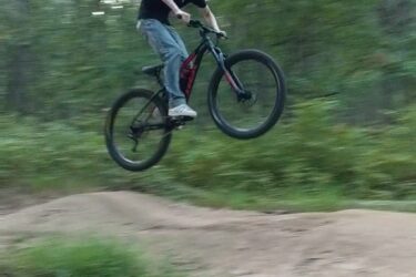 A person wearing a blue helmet is jumping off a dirt ramp on a mountain bike, with trees and greenery blurred in the background, suggesting high speed and action in a forested area. Frost Woods mountain bike trail.
