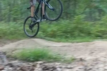 A person wearing a helmet and a green shirt is performing a jump on a mountain bike, catching air above a dirt ramp in a wooded area. The background features lush greenery, indicating a forest trail. Frost Woods mountain bike trail.