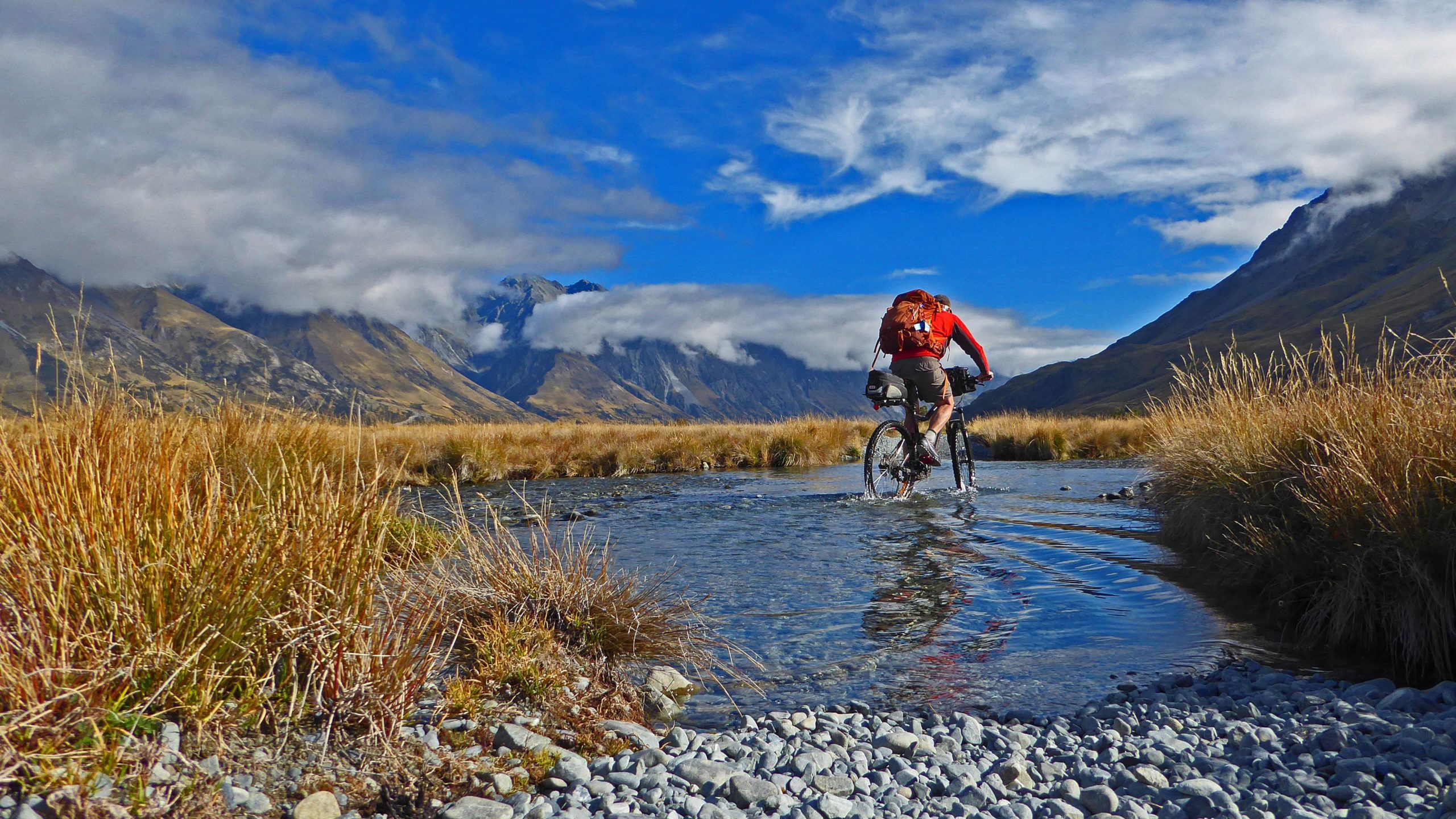 A cyclist riding through a shallow stream in a mountainous landscape, with rugged hills and valleys in the background under a blue sky filled with clouds. The scene features golden grasses and scattered pebbles along the water's edge. Godley Valley mountain bike trail.