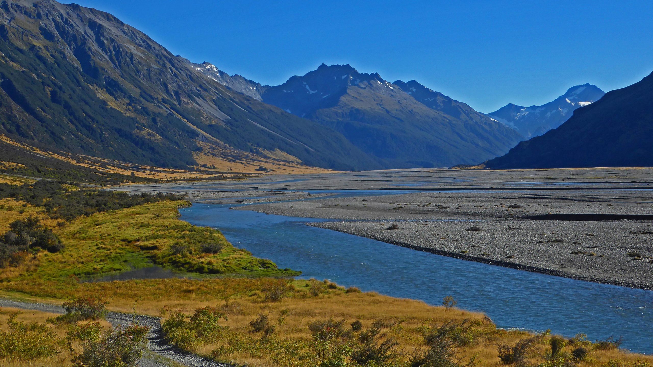 A scenic landscape featuring a winding river flowing through a valley surrounded by mountains. The foreground includes lush green vegetation and gravel paths, while the mountains in the background are partially covered with snow under a clear blue sky. Hopkins Valley to Monument Hut mountain bike trail.