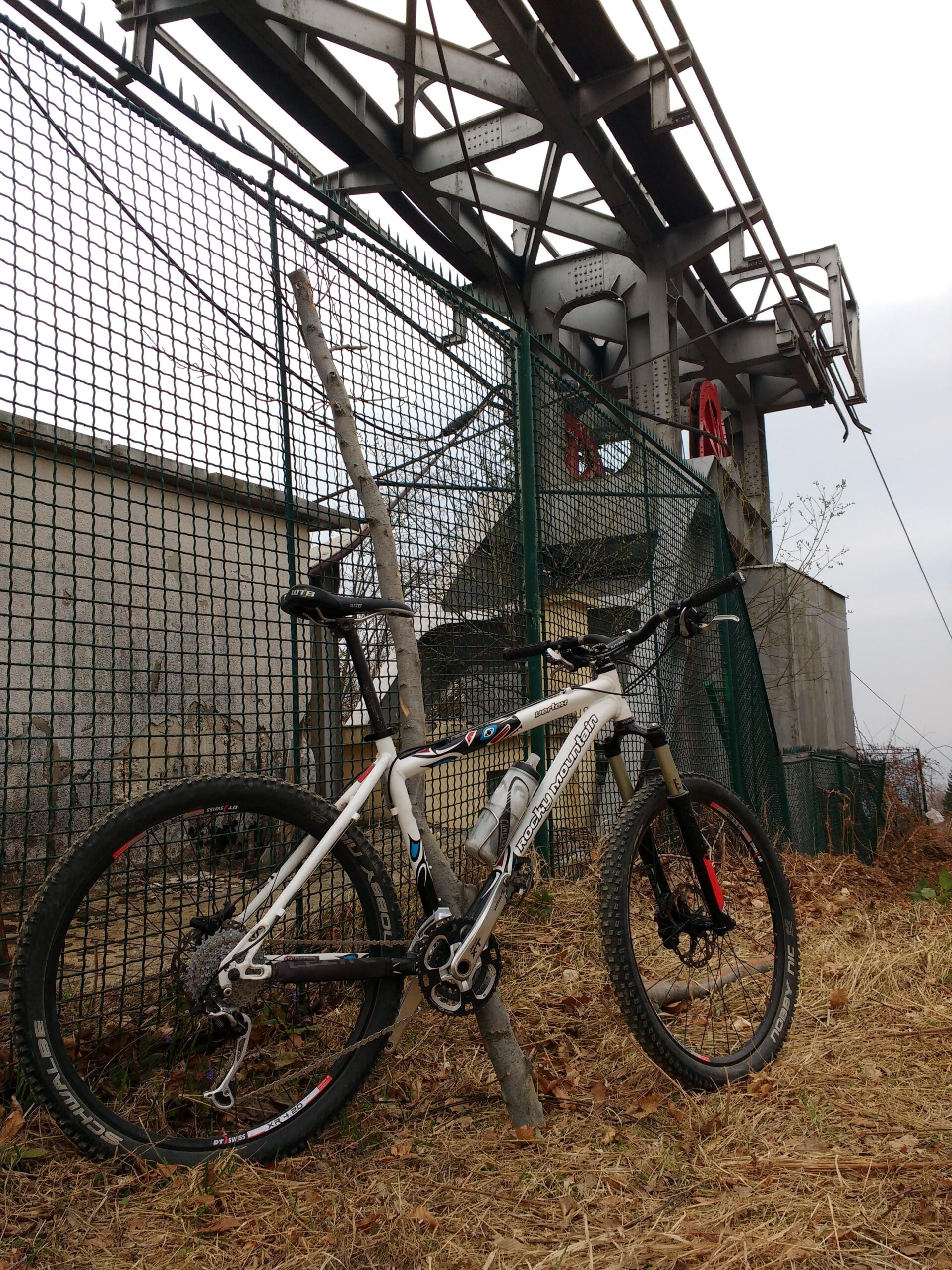 Rocky Mountain Vertex SE: A mountain bike leaning against a wooden post near a green fence, with industrial structures and a cloudy sky in the background. The area appears to be a mix of grass and dry leaves.