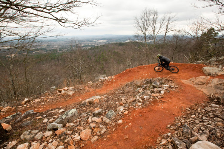 Mountain biker navigating a winding dirt trail on a hillside, surrounded by rocky terrain and bare trees, with an expansive view of the valley below on a cloudy day.