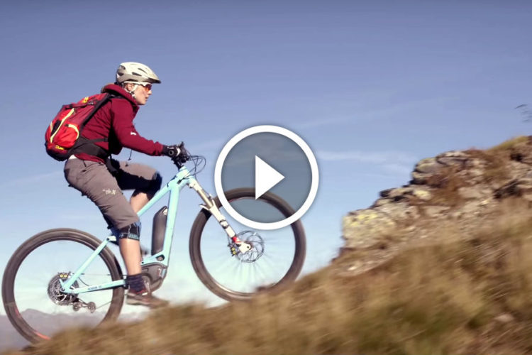 A mountain biker ascends a rocky slope on a sunny day, wearing a helmet and a backpack. The cyclist is clad in a maroon top and shorts, showcasing an action-packed moment against a backdrop of scenic mountains and clear skies.
