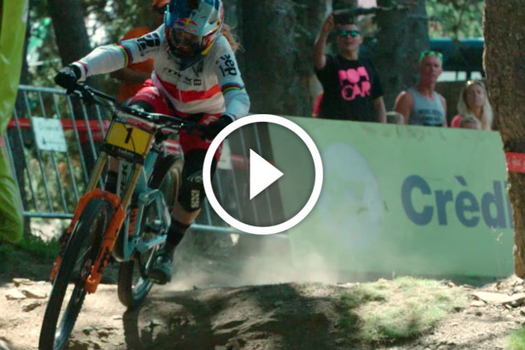 A mountain biker races down a rocky trail, kicking up dust as they navigate a challenging section of the course. They are wearing a helmet, colorful jersey, and riding a bike with the number 7 displayed on its frame. Spectators can be seen in the background, cheering and enjoying the event. The scene captures the excitement of downhill racing in a lush, wooded environment.