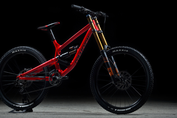 A red mountain bike with a sleek frame and high-performance suspension, positioned on a black background. The bike features prominent Maxxis tires and gold and black suspension components, highlighting its design for rugged terrain and advanced riding.